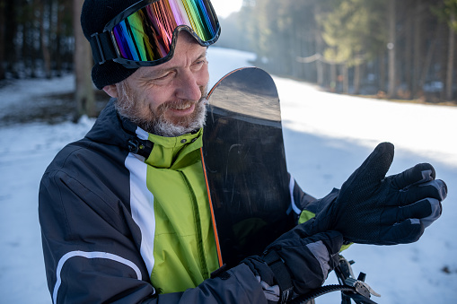 A cheerful elderly man with gray hair and a beard wearing a sports ski suit in green, black, gray, and white. On his head, he wears a black knitted hat and ski goggles. He adjusted his gloves. His snowboard rests against his chest. A sunlit ski slope can be seen in the background.