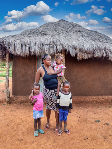 Happy village african family, mother with braided hair together with the three kids in front of the mud house with thatched roof