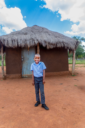 village african kid, in school uniform standing in front of the mud house shack in the yard