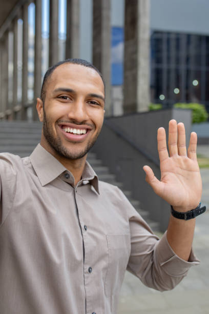 istock Hispanic smiling young man standing near office building and talking on camera phone, greeting and waving. Vertical photo 2028487790