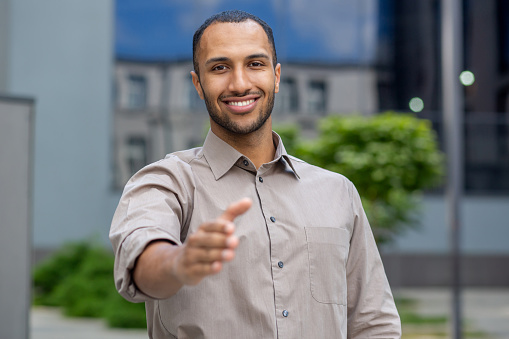 Portrait of a successful and smiling young businessman, hispanic man standing outside an office building and extending his hand to the camera in greeting.