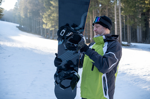 A senior man with graying hair and a beard carefully examines his board. He is dressed in a green, black, gray, and white sports ski suit. On his head, he wears a black knitted hat and ski goggles. In the background, a sunlit ski slope is visible.