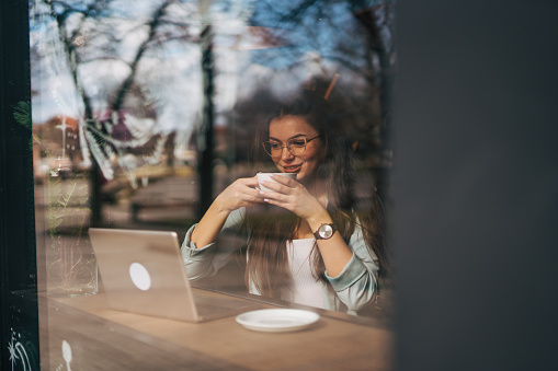 A young woman uses her laptop and does online work in a coffee shop while drinking coffee.