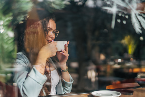 A young woman is sitting in a cafe by the window drinking coffee and using her phone.