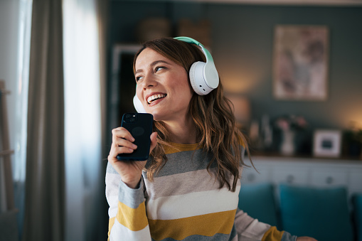 Shot of young woman using smartphone and headphones in living room and dancing