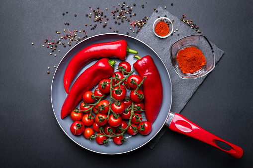 Ceramic pan full of fresh vegetables in red color: tomatoes and peppers. Kitchenware with a red handle on the dark background with spices around.