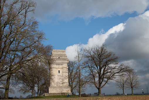 The Bismarck Tower near Augsburg stands between bare trees in February. A cyclist rides past it. The sky is blue with white clouds.