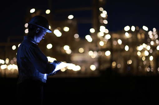 male engineer with a reflective safety jacket works at late night with the bokeh from factory lights