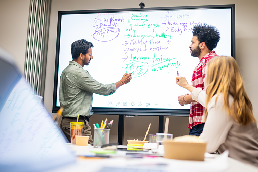 Two young businessmen and their diverse team brainstorming on a interactive whiteboard in a modern office.