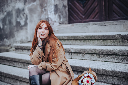 Fashion portrait of a stylish red hair woman in coat sitting on old stairs on city street downtown with flowers next to her. Fashionable young woman is posing on rustic stairs and looking at camera.