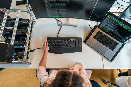 Top view of a female software engineer coding on dual monitors in a contemporary office. She is dressed in casual attire, focusing intently on a complex programming task. Her workstation is equipped with specialized hardware, reflecting a professional tech setting.