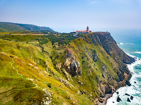A red and white lighthouse in Cabo Da Roca perched on a rugged cliff. The lighthouse stands against a clear blue sky, its beacon reaching out over the vast ocean. Surrounding the cliff, lush greenery and walking trails add to the scenic coastal landscape.
