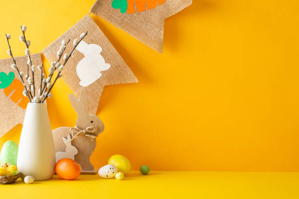 Easter theme. Side view shot of table with rustic elements, vase with plant, wooden rabbit figures, colored eggs, nest holding chick against orange backdrop, highlighted by string of festive flags