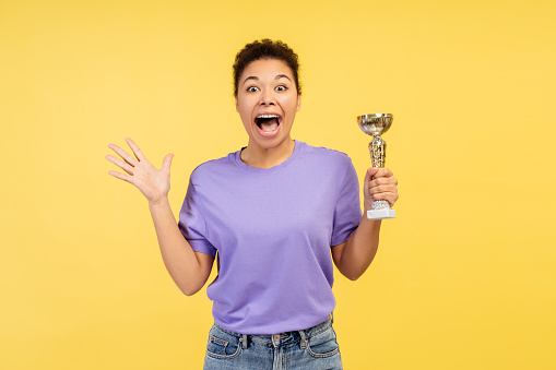 Excited smiling African American woman, competition winner holding trophy cup looking at camera standing isolated on yellow background. Concept of victory, sport