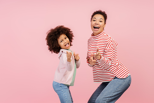 Smiling, excited African American woman, and little cute girl showing fingers looking at camera isolated on pink background. Concept of shopping, sale