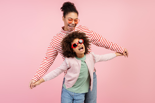 Portrait of smiling beautiful mother and little cute daughter wearing stylish sunglasses dancing isolated on pink background. Concept of fun, motherhood, childhood