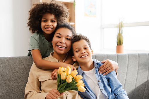 Happy African American family, little daughter and son hugging beautiful mother, holding bouquet of yellow tulips sitting on sofa at home, looking at camera. Concept of holiday, birthday, Mother's day