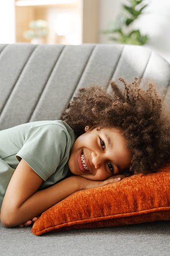 Smiling little cute girl lying on pillow on comfortable sofa looking at camera at home. Childhood concept, advertisement