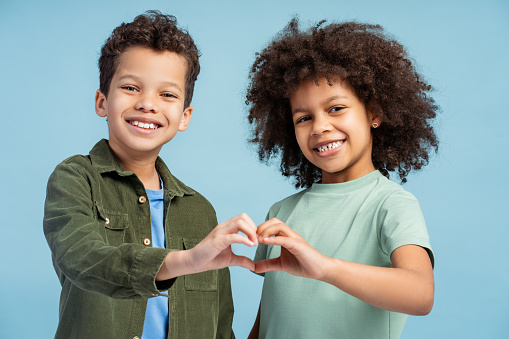 Smiling African American boy and girl, positive sister and brother gesturing, showing heart with hands looking at camera isolated on blue background. Concept of love, family