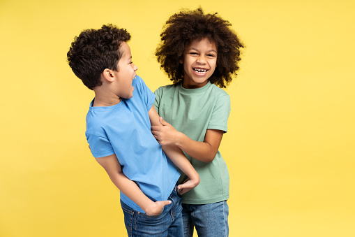 Beautiful, smiling children, African American little boy and girl fooling around together, looking at camera posing isolated on yellow background. Concept of relationships, childhood, family