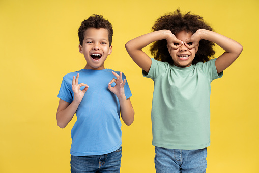 Portrait of positive, smiling African American boy and girl showing ok sign, gesturing looking at camera standing isolated on yellow background. Concept of fun, childhood