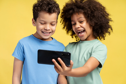 Happy beautiful African American friends, boy and girl holding mobile phone, using mobile app, watching video standing isolated on yellow background. Online technology concept