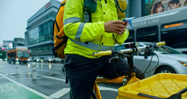 A faceless cargo bike delivery person looking at their phone while waiting with their bike on the streets of downtown Los Angeles on a rainy day in winter.