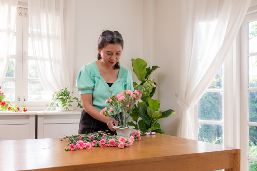 A happy Chinese retired woman in a green shirt arranging pink carnation flowers in a vase to add a decorative touch to is living room at home, relaxing during weekends