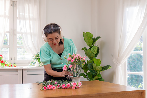 A happy Chinese retired woman in a green blouse  arranging pink carnation flowers in a vase to add a decorative touch in living room at home, while listening to music using wireless headphone, relaxing during weekends