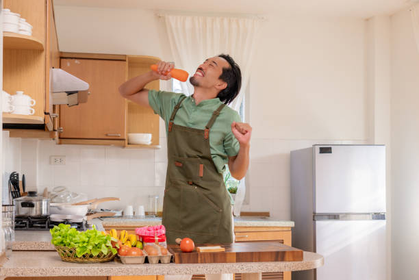 A funny Japanese mid-adult man dancing and singing in a kitchen while preparing his meals, using carrot as microphone, relaxing at home on weekends