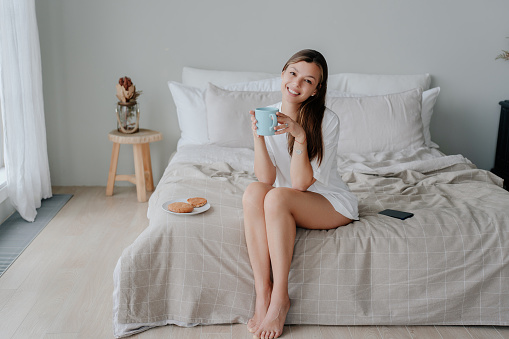 Smiling woman with coffee on bed, cookies, morning, relaxation, bedroom, cheerful, comfortable, serene, white dress, barefoot