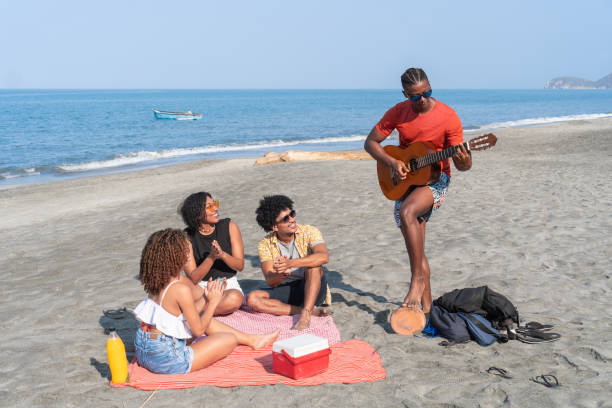 Friends gathered on beach blanket, enjoying guitar music and sunny beach vibes.