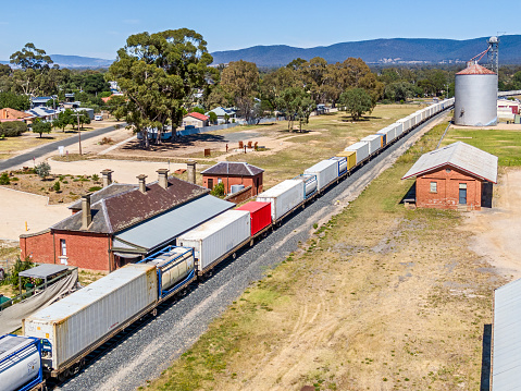 Aerial view container freight train passing heritage-listed railway station in pretty rural town with a backdrop of low hills. Grain silo next to railway tracks, some town residences visible, most shipping containers on the train are white with one red and one yellow visible. Logos & ID edited. (Avoca in Victoria's Pyrenees)