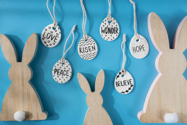 Group of wooden bunny ears and clay Easter eggs with words RISEN HOPE BELIEVE PEACE LOVE WONDER on blue background. Children air dry clay activity handicraft idea. Preparing for Easter holiday decorating. Modern organic design minimalistic plastic free sustainable decor