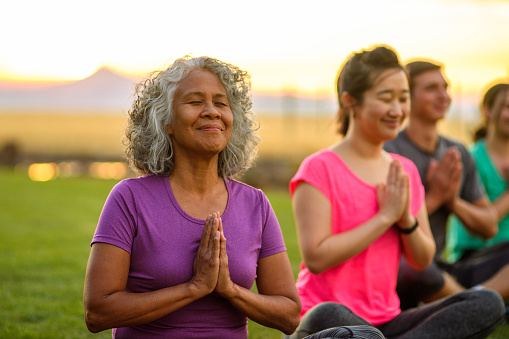 An active senior woman of Hawaiian and Chinese descent meditates with her eyes closed while attending an group fitness class outdoors at a public park on a beautiful summer evening.