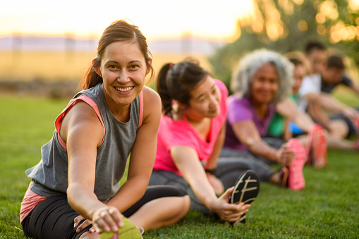 Portrait of a fit woman smiling directly at the camera while doing a seated leg stretch during a group fitness class outdoors in a public park on a beautiful summer evening in Oregon.