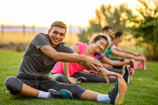 Portrait of a fit young man smiling directly at the camera while doing a seated leg stretch during a group fitness class outdoors in a public park on a beautiful summer evening in Oregon.
