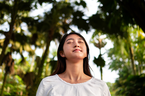 Front view of an Asian woman with eyes closed and head raised enjoying morning breeze in the nature.