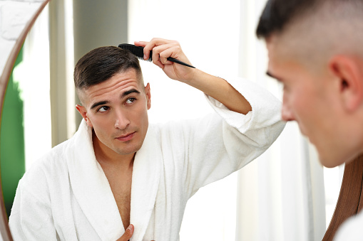 Handsome young man combing his hair while standing against mirror in the bathroom close up