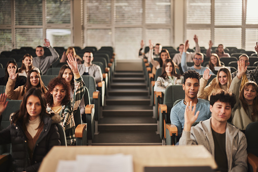 Large group of happy college students raising their hands to ask a question during a class in amphitheater and looking at camera.