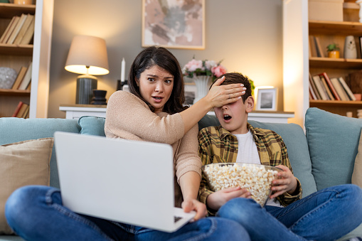 In their living room, a mother and her son share a thrilling movie night. With popcorn in hand, they watch a shocking film on the laptop, mother covering sons eyes with hand