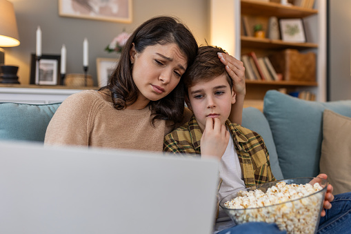In their living room, a mother and her son share a cozy movie night. With popcorn in hand, they watch a sad film on the laptop, snuggled up on the sofa, finding comfort in each other's company.