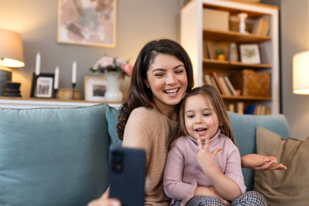 istock Smiling mother and daughter on video call using digital phone at home. while making video call and waving hello sitting on sofa. Woman and her cute little kid doing video conference with grandparents. 2029606586