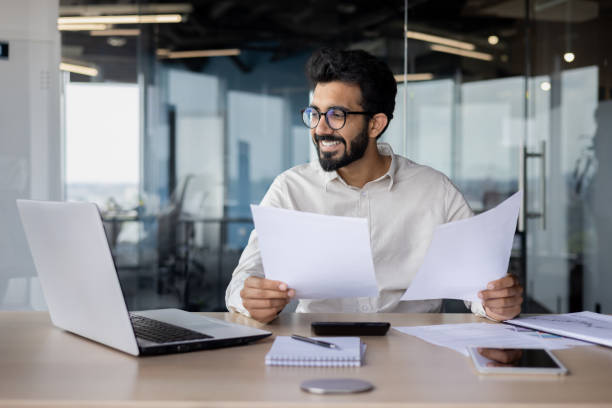 istock A young Indian man works in the office with documents, smilingly looks at a laptop, talks on a video call, holds bills and an agreement in his hands 2029608249