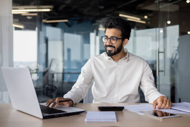 istock Smiling indian businessman working at a desk with laptop in modern office setting 2029608253
