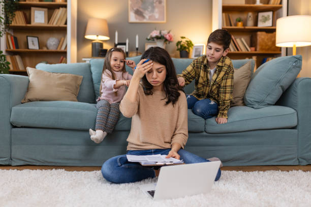 Tired young mother sitting on the floor and working with laptop and documents while little kids and jumping on the sofa and having fun and making noise
