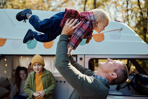 Playful father having fun with his baby boy during autumn day at trailer park. Their family is in the background.