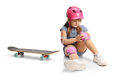 Girl with skateboard, wearing knee pads and helmet and sitting on the floor with injured knee isolated on white background