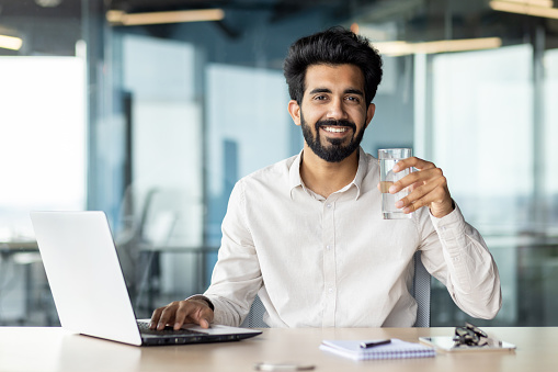 An Indian young male businessman is sitting in the office at his desk and holding a glass of clean water in his hand. He looks at the camera with a smile.