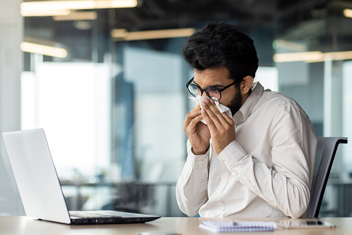 A sick young Indian man is not feeling well at the workplace. He sits at the desk in the office and wipes his nose with a napkin from a runny nose.